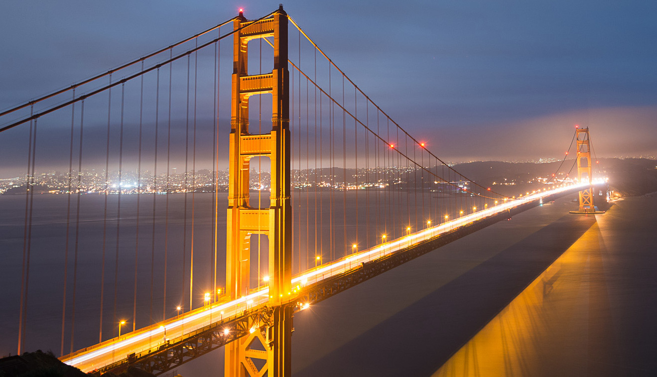 A photo of the Golden Gate Bridge at dusk with car headlights appearing as a solid stream of light on the bridge